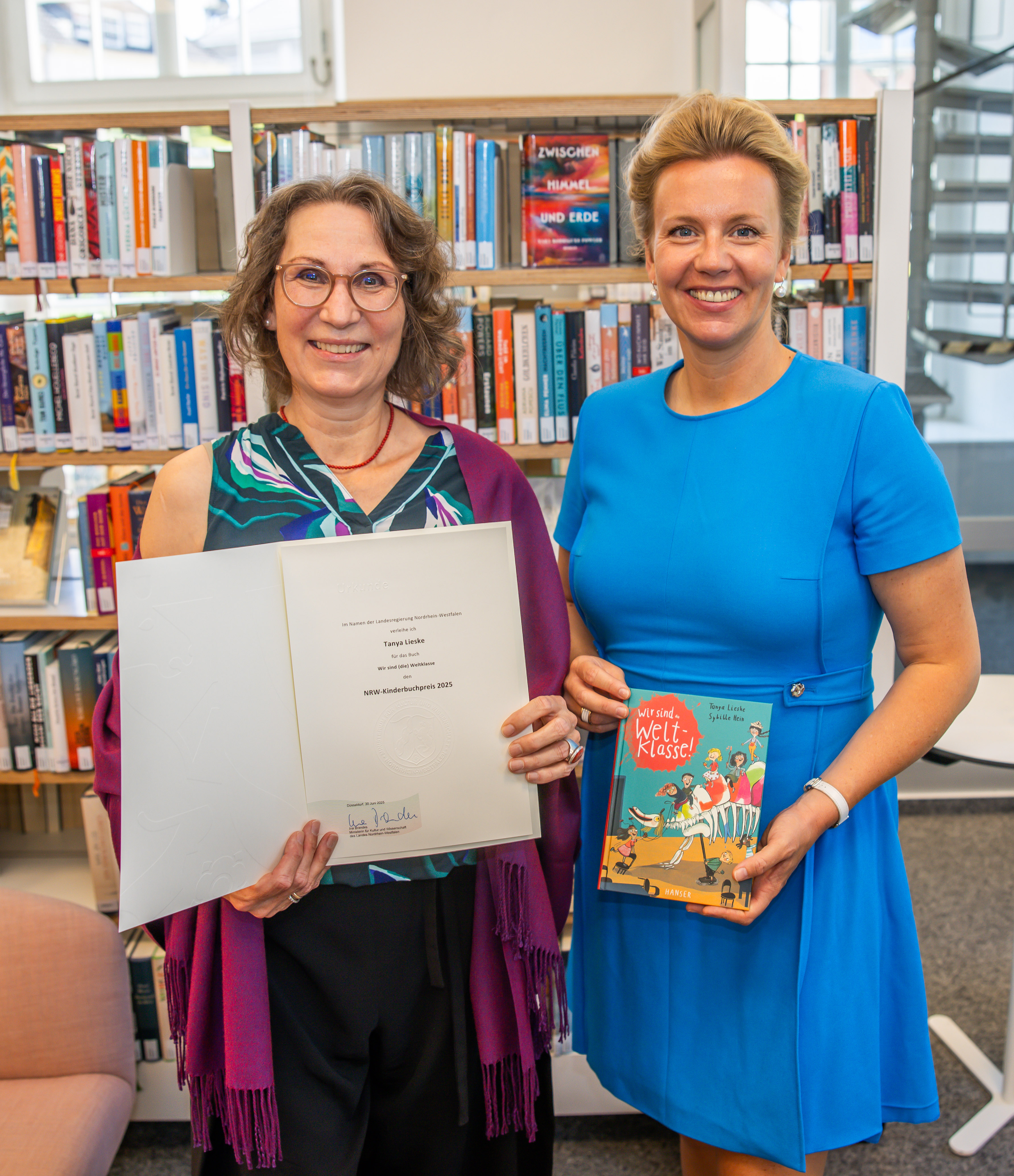 Zwei Frauen lächeln in die Kamera mit einer Urkunde und einem Buch in der Hand