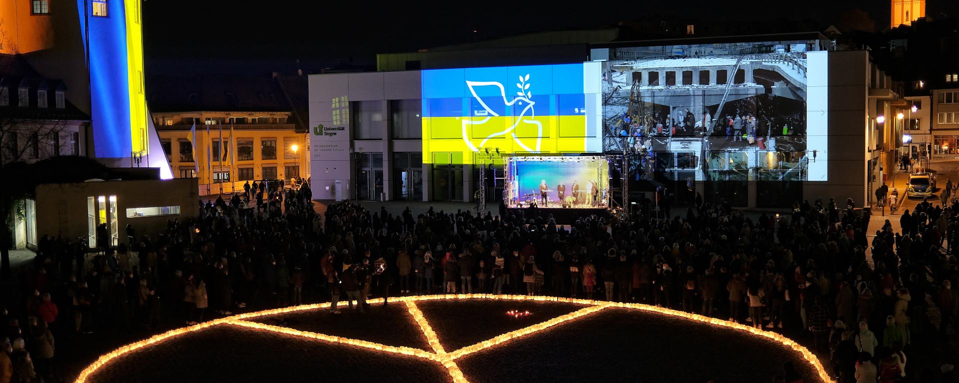 Ein aus Kerzen gelegtes Friedenssymbol auf einem Vorplatz mit der ukrainischen Flagge im Hintergrund