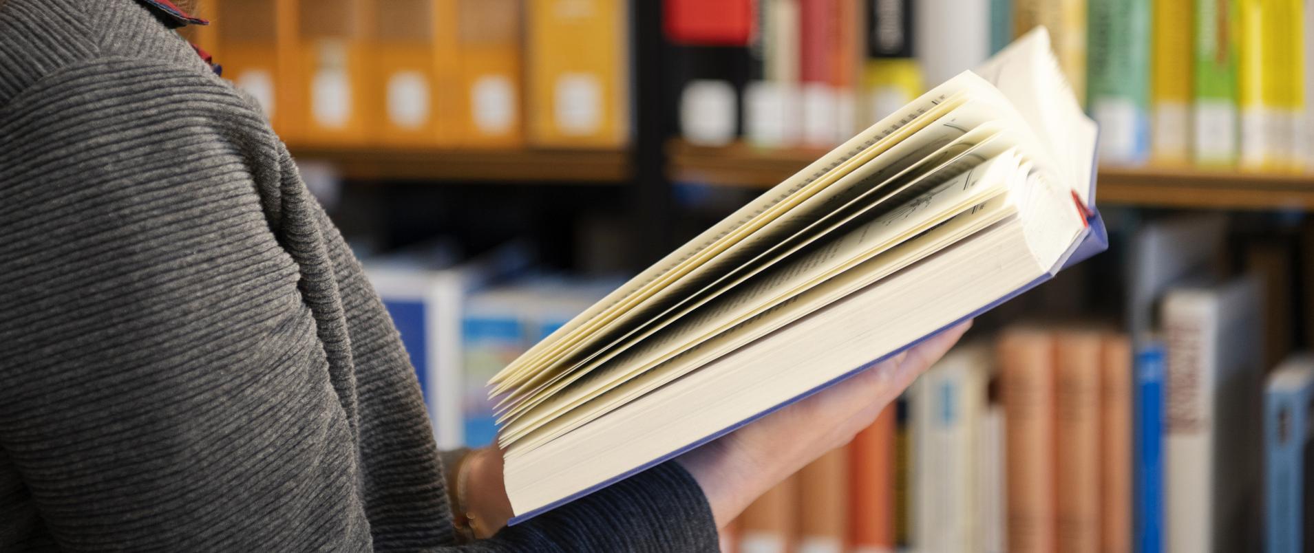 Eine Frau steht vor einer Bücherwand und blättert durch ein Buch.