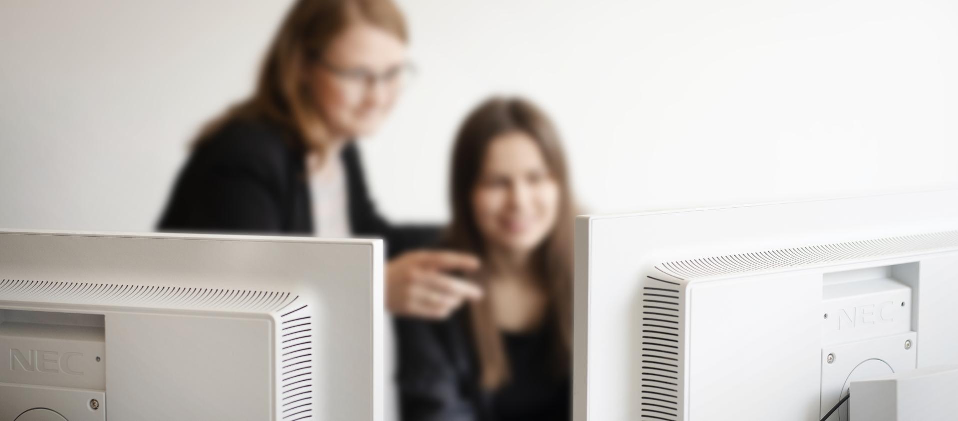 Zwei Computerbildschirme im Vordergrund, im Hintergrund zwei Frauen, die damit interagieren