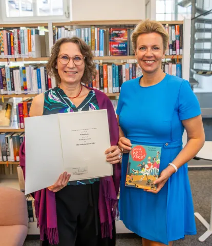 Zwei Frauen lächeln in die Kamera mit einer Urkunde und einem Buch in der Hand