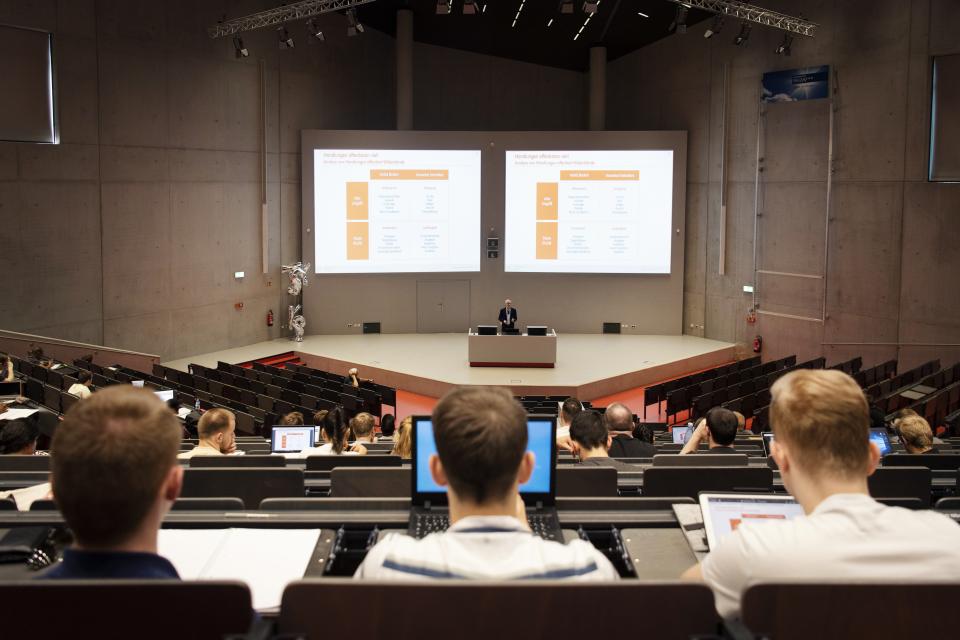 In einem Hörsaal schauen drei junge Menschen nach vorne auf den Vortrag des Dozierenden.