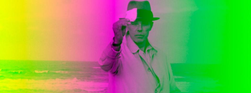 Foto von Joseph Beuys, er hält etwas in die Höhe. Im Hintergrund das Meer und Strand, verfremdet durch die Überlagerung mit verschiedenen Neonfarben