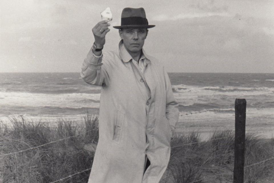 Foto von Joseph Beuys, er hält etwas in die Höhe. Im Hintergrund das Meer und Strand