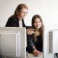 Zwei Computerbildschirme im Vordergrund, im Hintergrund zwei Frauen, die damit interagieren